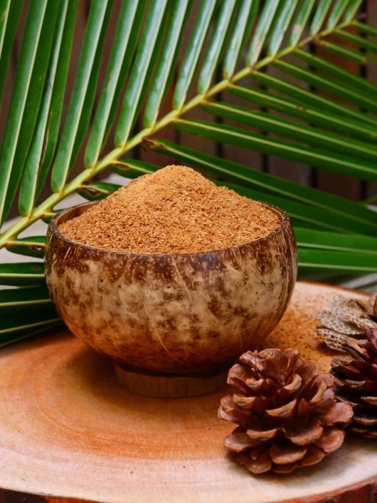 A rustic wooden bowl filled with organic coconut sugar, surrounded by pinecones and tropical foliage on a wooden surface.
