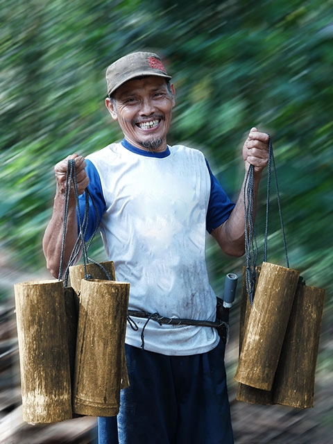 A smiling Realsa farmer holding wooden sap containers in a tropical forest, showcasing traditional coconut sugar harvesting.