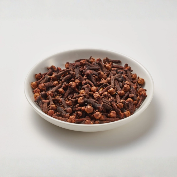 A top-down view of a small white ceramic bowl filled with a large amount of dried, whole clove buds, showcasing their signature dark reddish-brown color against a plain white background.