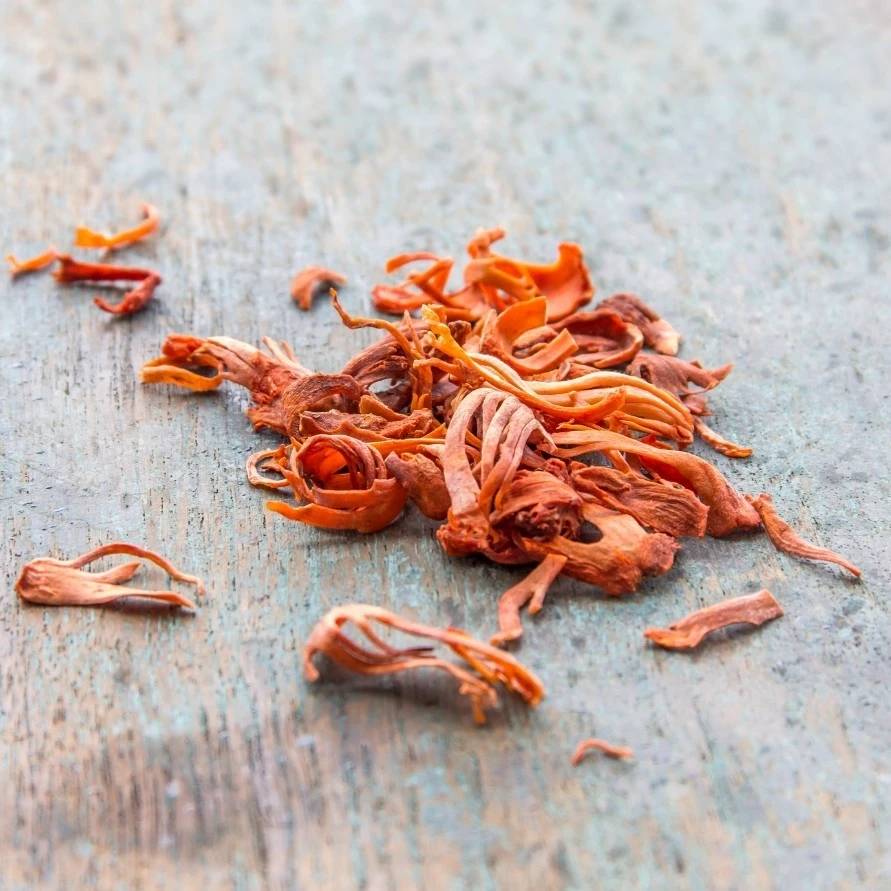 A close-up photograph of a small pile of dried, bright orange-red mace blades centered on a rustic, weathered light-blue painted wooden surface with a few scattered fragments.