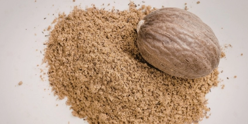 A top-down, close-up photograph of a whole brown nutmeg seed resting next to a large pile of finely ground nutmeg powder on a clean, light-colored surface.