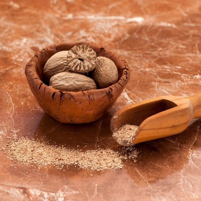 A detailed close-up photograph of whole nutmeg seeds and a halved seed in a small, rustic clay bowl, positioned next to ground nutmeg powder in a wooden scoop and sprinkled on a polished brown marble surface.
