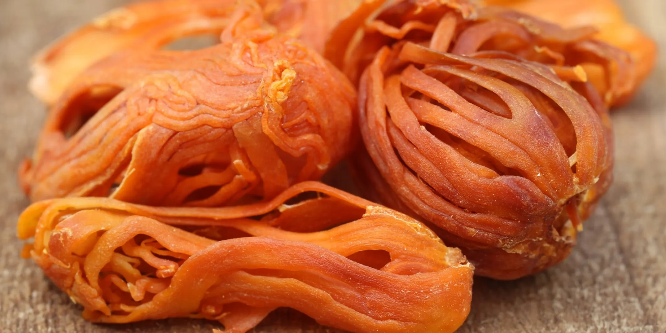 A detailed macro photograph showing whole, dried mace blades piled on a textured wooden surface. The vibrant orange-red color and intricate, web-like lace structure of the mace are clearly visible under focused light.