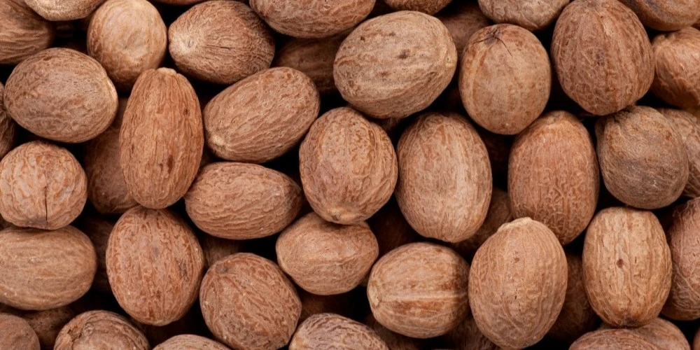 A high-resolution, top-down close-up photograph of a dense pile of whole, textured nutmeg seeds showcasing their characteristic brown color and wrinkled surfaces.