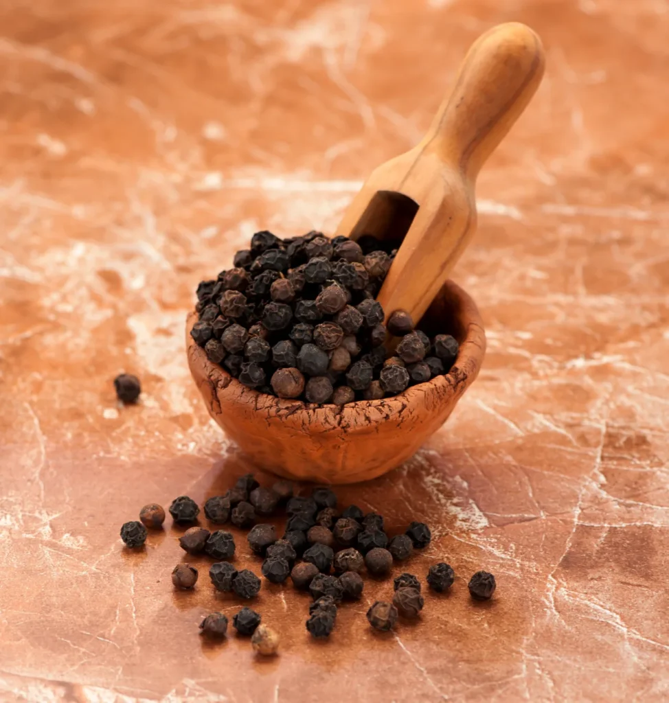 A close-up photograph of whole black pepper peppercorns piled inside a small, rustic terracotta clay bowl next to a wooden spice scoop, with several peppercorns scattered on a polished brown marble surface in the foreground.