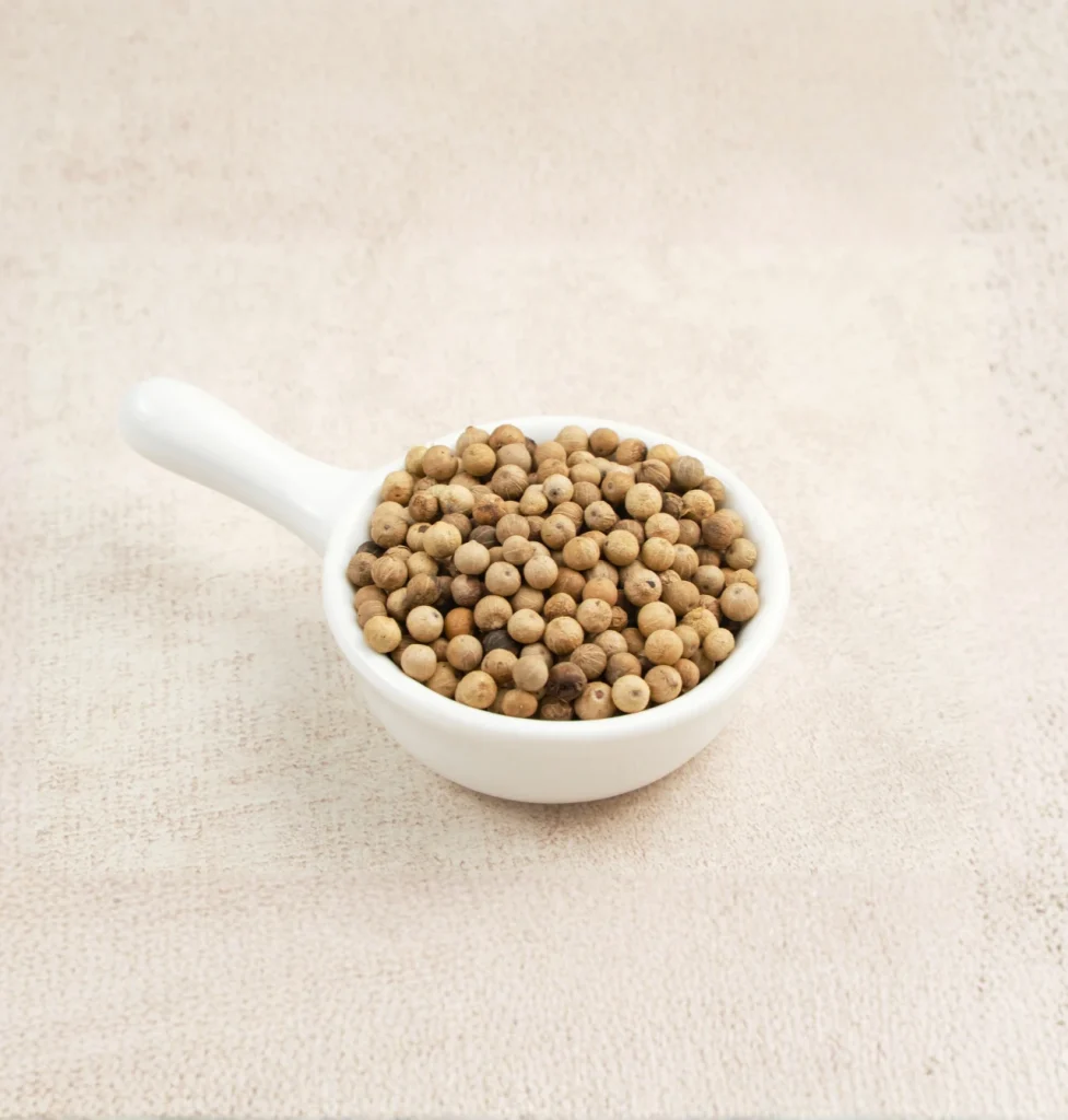 A top-down, minimalist photograph of a small white ceramic bowl with a handle filled to the brim with whole white pepper peppercorns, centered on a light beige, textured background.