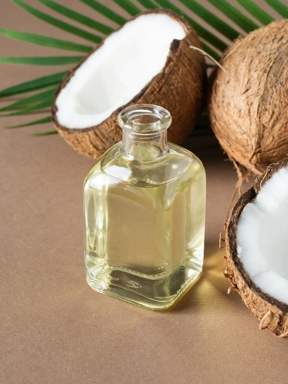 RBD coconut oil in a clear glass bottle beside fresh halved coconuts and a green palm leaf