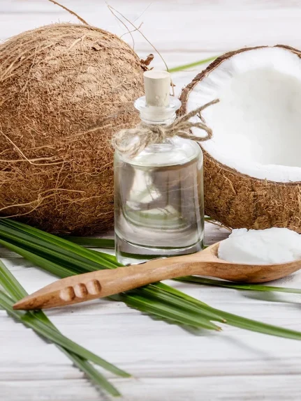 A bottle of virgin coconut oil with whole and halved coconuts, coconut cream, and palm leaves on a white wooden surface.